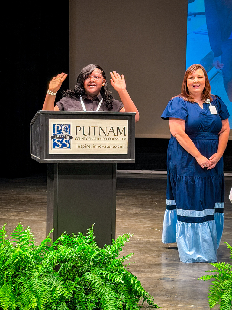 Woman at podium waves with two hands raised, another woman stands nearby, both wearing blue dresses.
