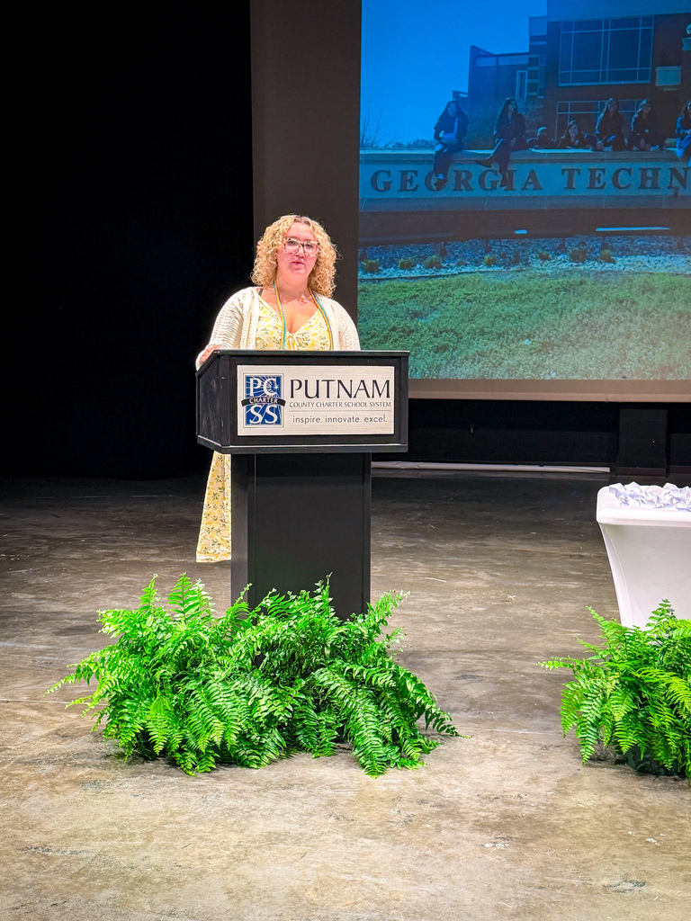 A woman stands behind a podium with a sign reading "PC Putnam SS" on a stage with green plants and a projector screen in the background.
