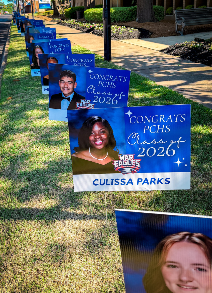 A row of signs on the grass, each featuring a photo of a person and the words "CONGRATS" and "2026".