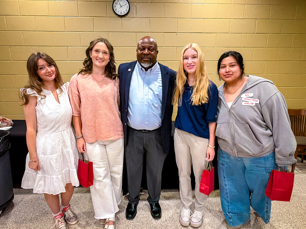 Five people stand in a row for a picture, all smiling. The man wears a navy blazer. Three women hold red bags.