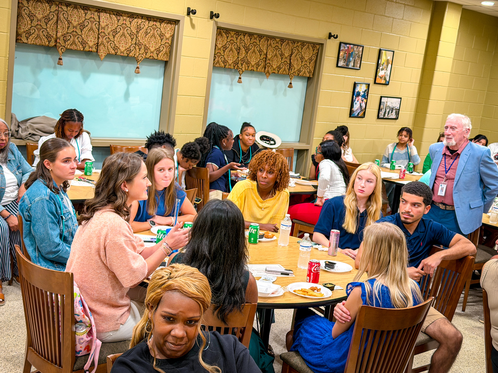 People in a room with yellow walls and windows, gathered around tables, some engaged in conversation.