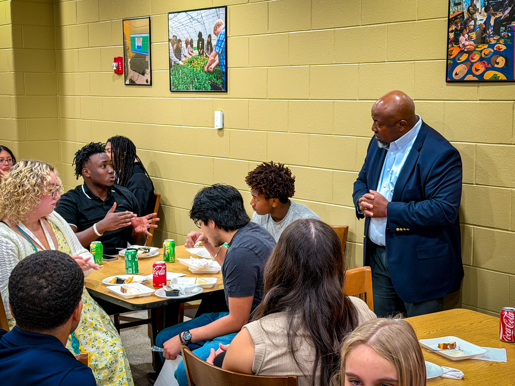 A group of people seated at tables in a room. A man stands in the background speaking. Artwork adorns the walls.