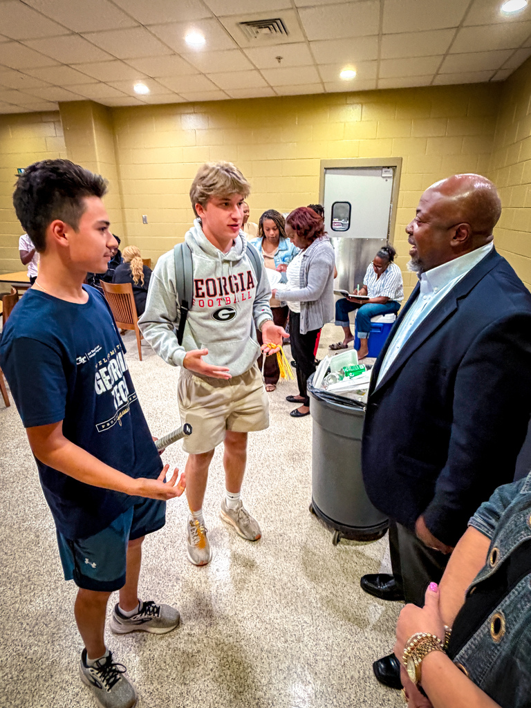 People stand in a brightly lit room, one in a business suit talks with two others; a trash can is behind them.