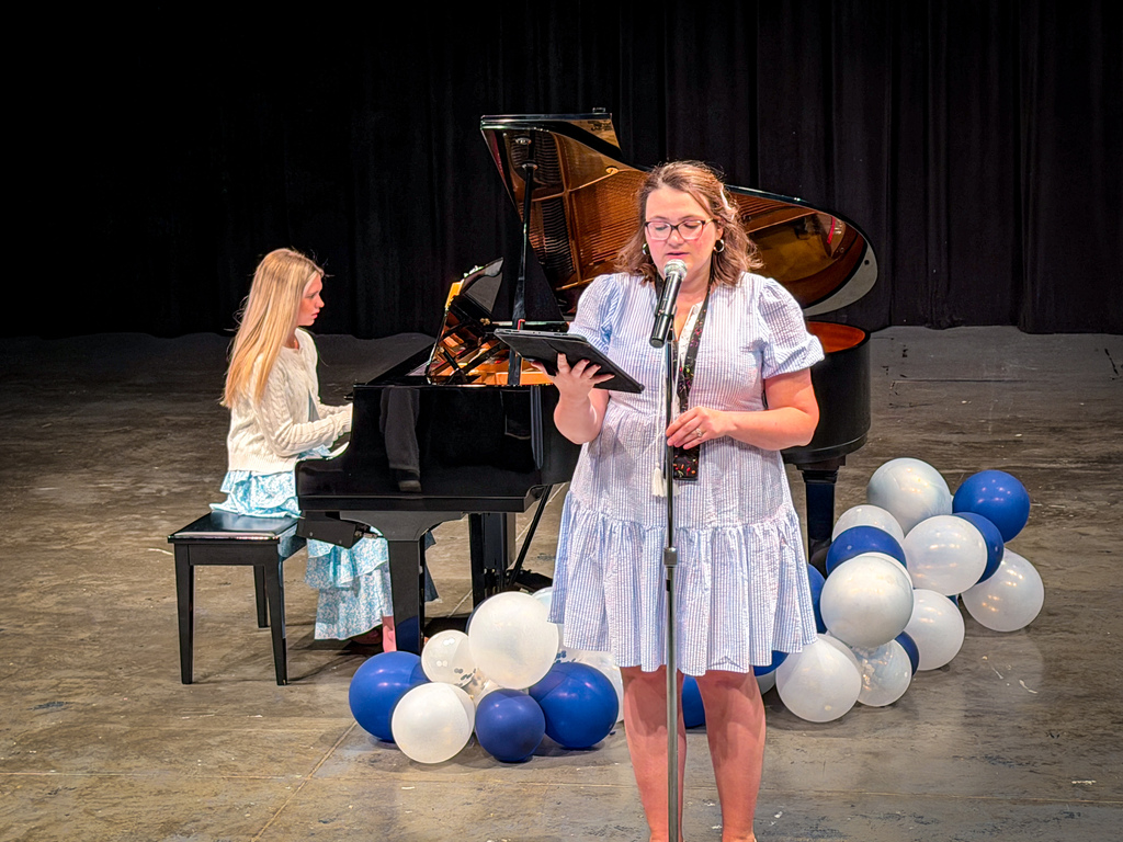 A woman stands with a microphone on a stage, reading from a tablet. Behind her, a piano is on a stage with a curtain backdrop.