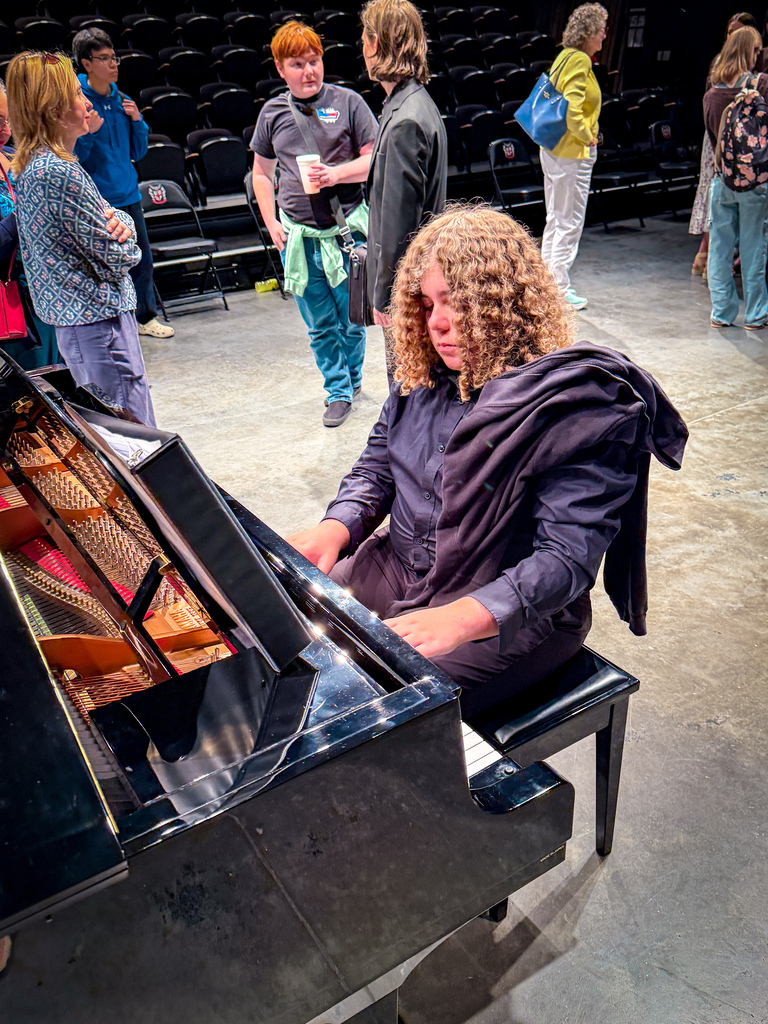 A person seated at a black piano in a theater lobby, with an audience standing and conversing in the background.