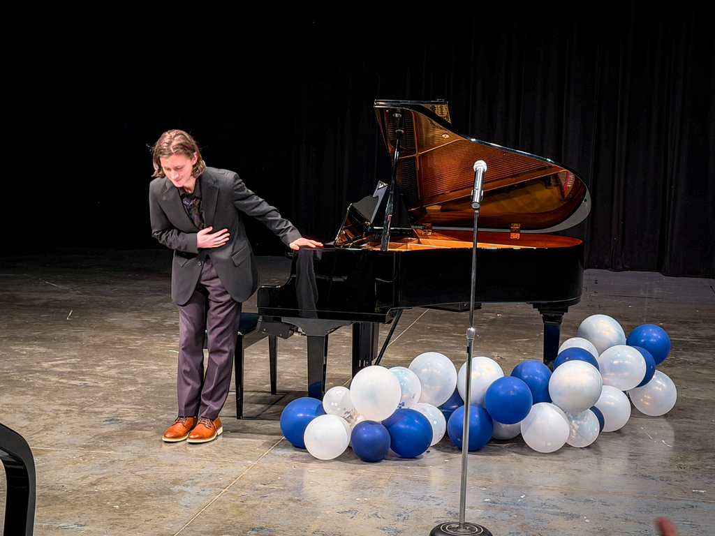 Person in a suit standing by a grand piano with a microphone. Many balloons are on the floor in front of the piano.