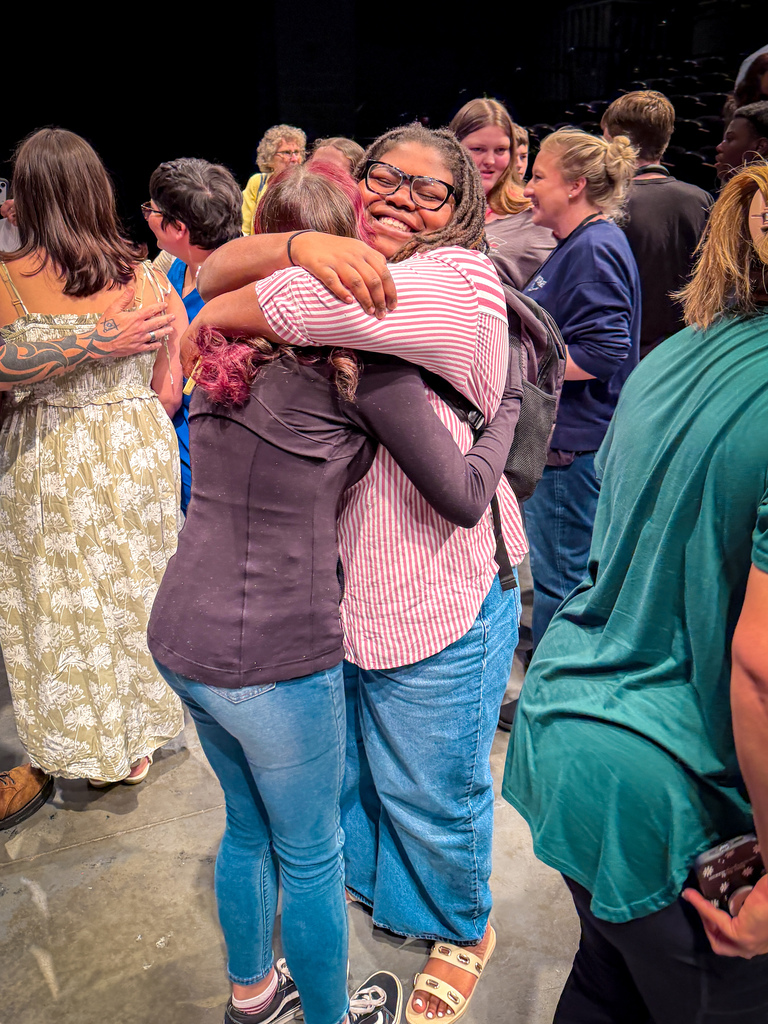 Several people in a room, two women hug each other in the center, others look on.