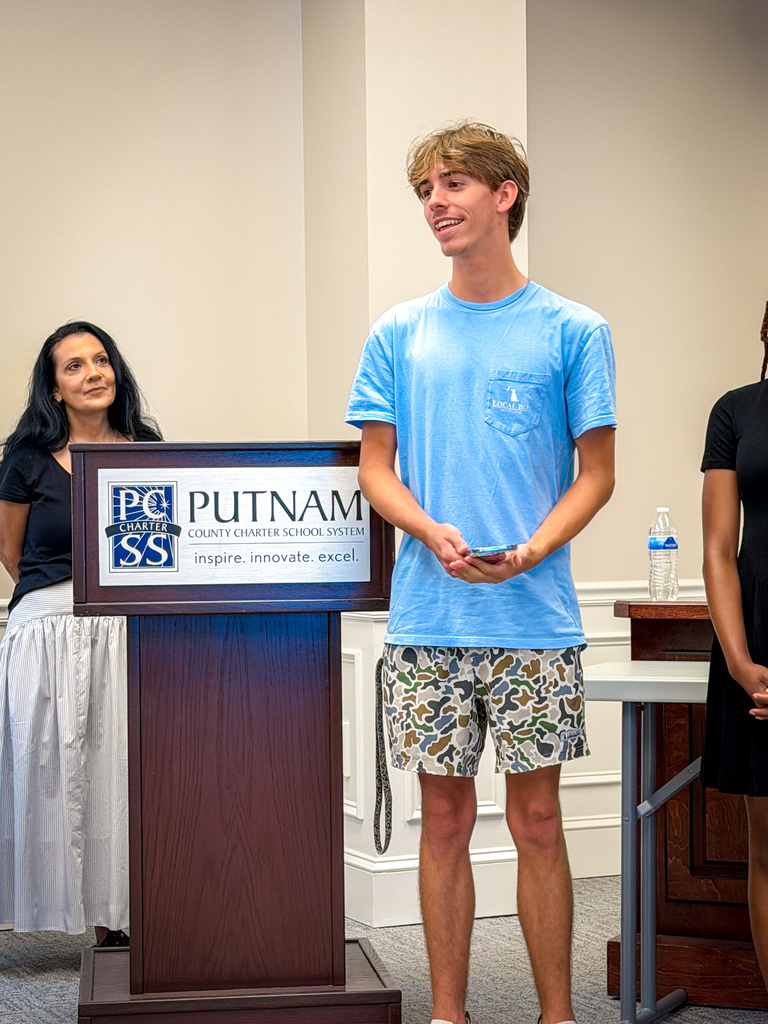 A young man in a light blue shirt and patterned shorts stands at a podium, smiling. Behind him, a woman in a white skirt looks at him.