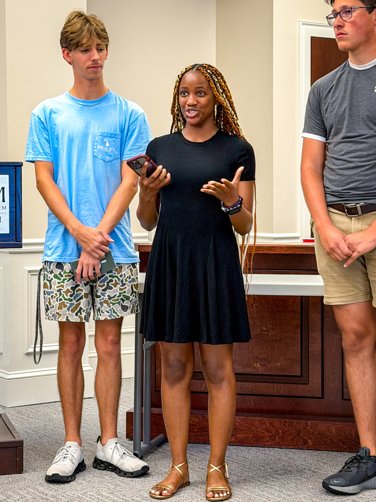 Three individuals in a room. A woman in a black dress speaks into a microphone. Two men stand behind her.