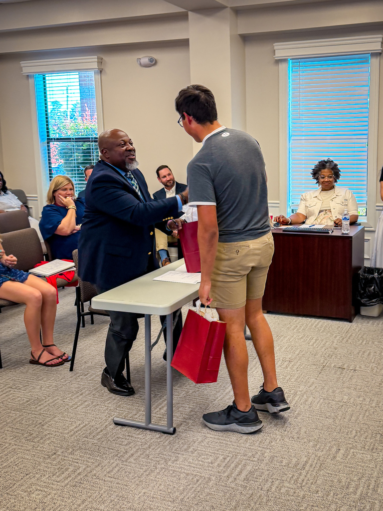 Man in suit hands red gift bag to man in gray shirt and khaki shorts in a room.