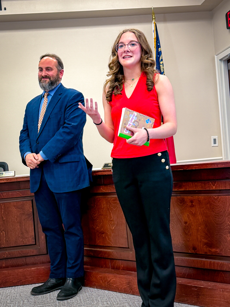 A man and a woman stand at a podium. The woman smiles and speaks while holding a book.