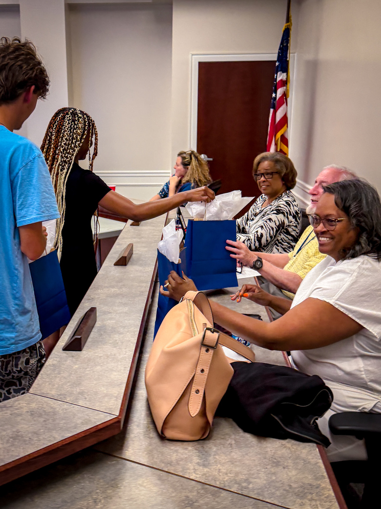People at a table receiving bags. A woman smiles while another hands her a bag. An American flag hangs on a wall.
