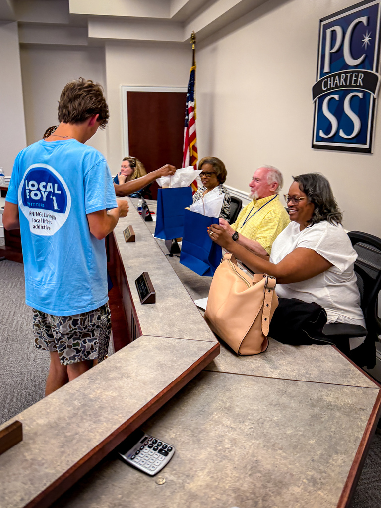 People at a meeting; a boy in a blue shirt hands out bags while others watch.