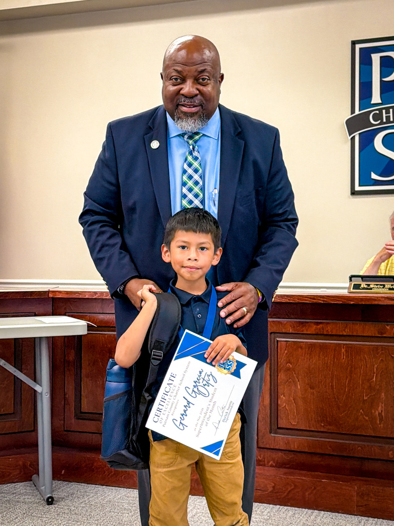 Man in business attire holding certificate, stands behind child in school uniform holding certificate, in front of counter.