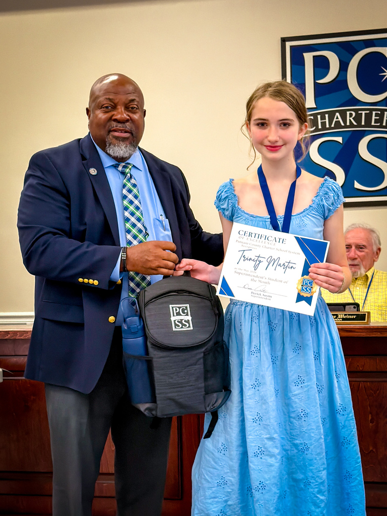 A man and girl stand together in a room holding a certificate. The man wears a suit, the girl wears a dress and a medal.