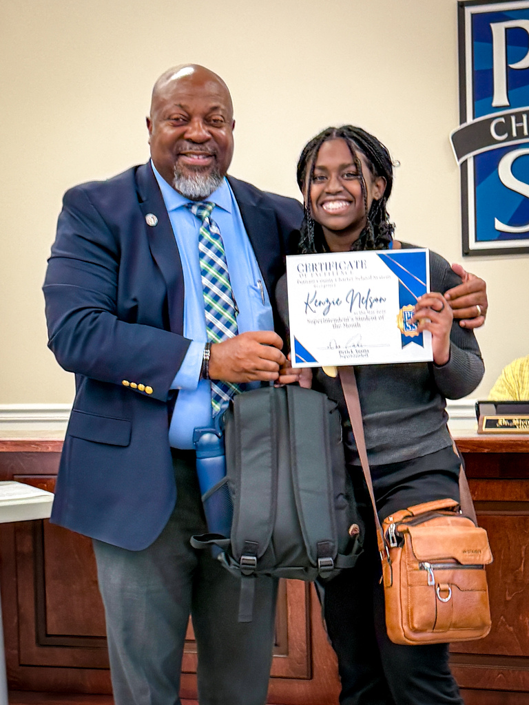 Man and woman stand together, smiling, holding a certificate and bags in front of a sign.