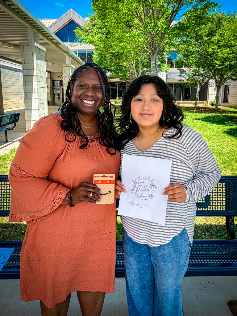 Two women stand next to a bench, smiling. One holds a paper and card; the other wears an orange dress. Behind them, a grassy area and trees.