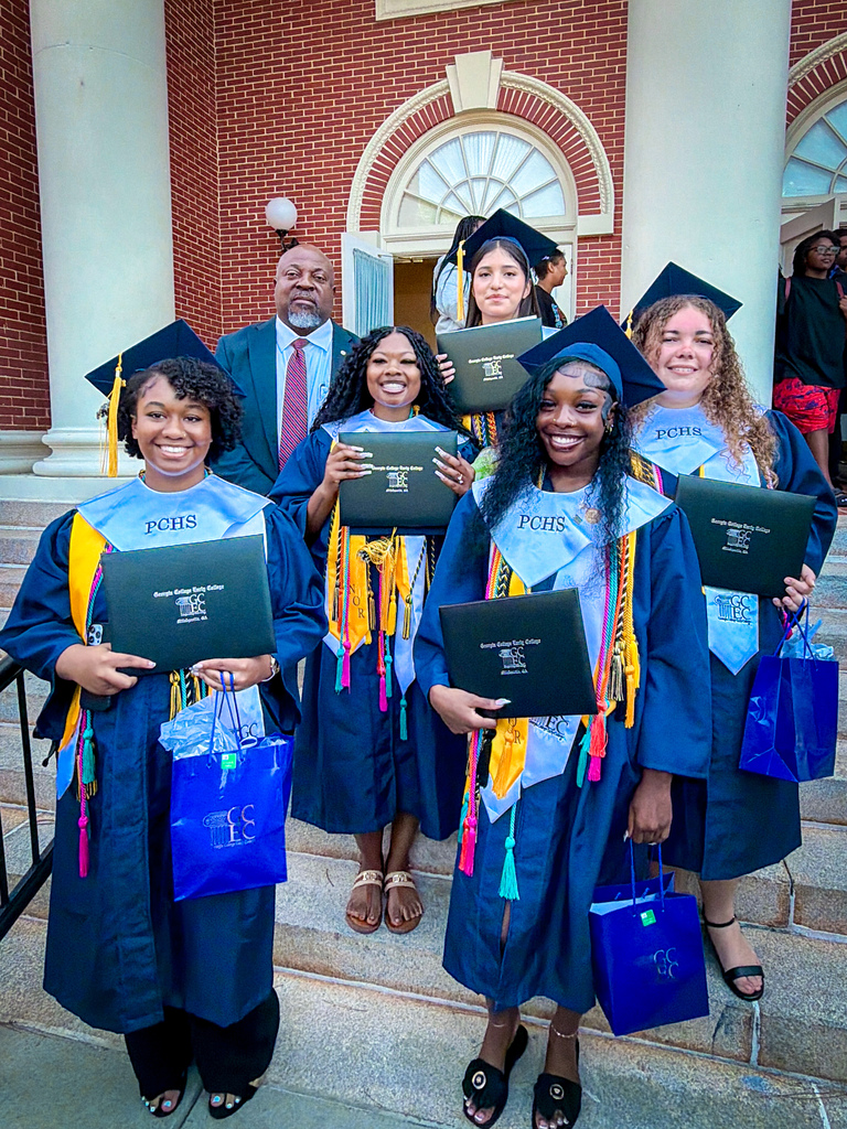 Graduates in blue gowns with yellow tassels pose on a brick-walled staircase with a man.