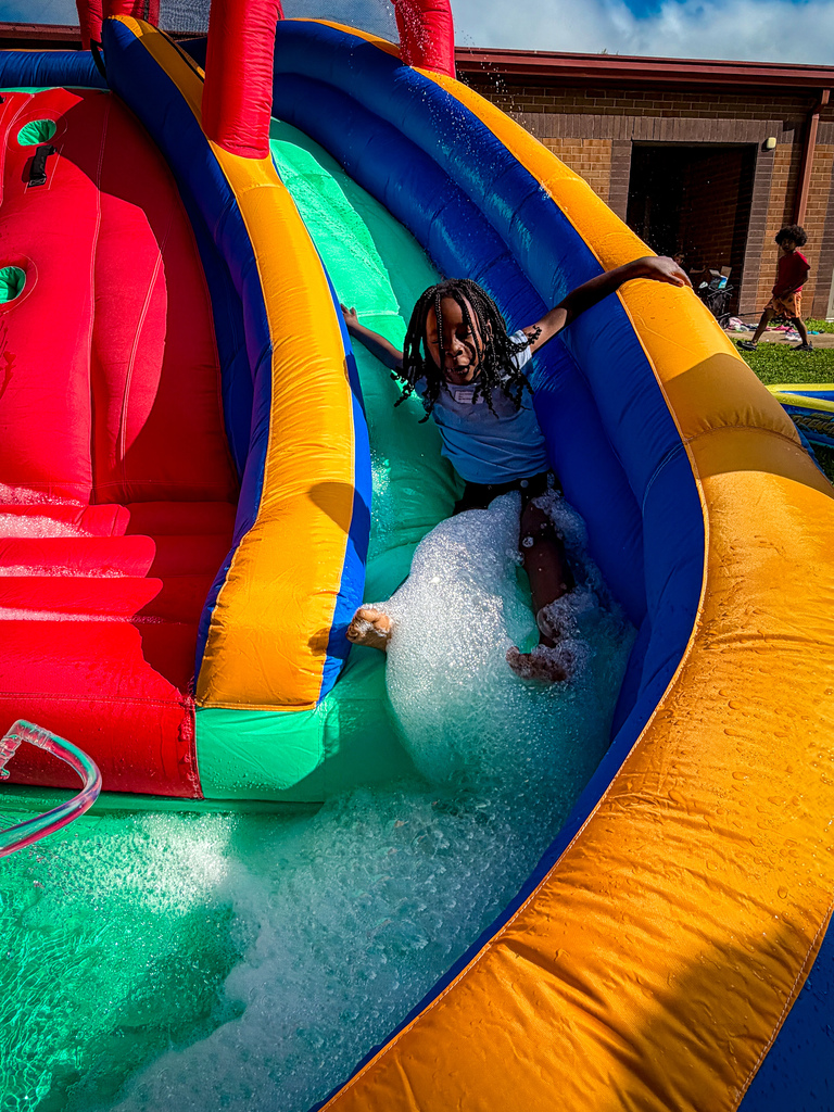 A child slides down a colorful, inflatable water slide with foamy water. Background shows a building and another child walking.