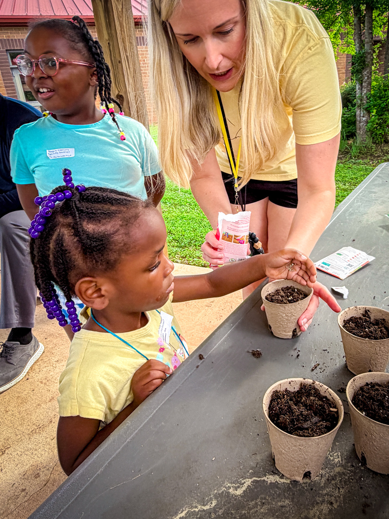 A woman helps a young girl plant seedlings into small pots. Three small plants in pots are already planted.