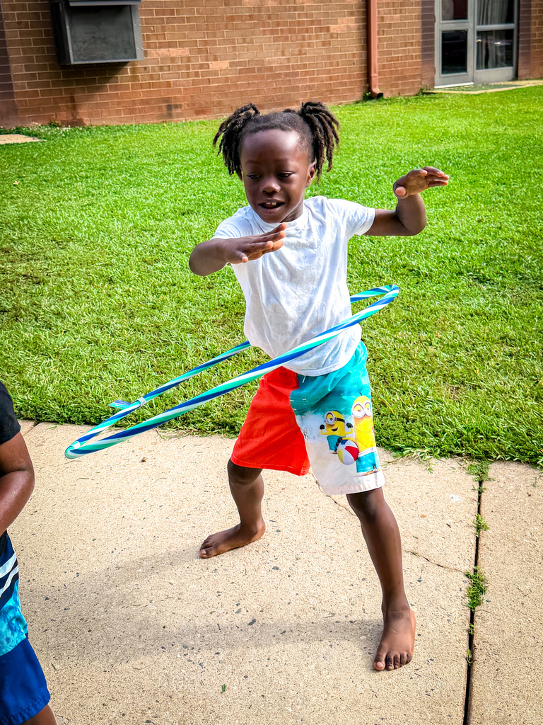 A child in a white shirt and multicolored shorts hula-hoops on a sidewalk, with a grassy area and building in the background.
