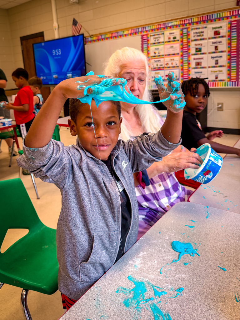 A young boy with blue paint on his hands raises them up. A woman helps him while other children are in the background.