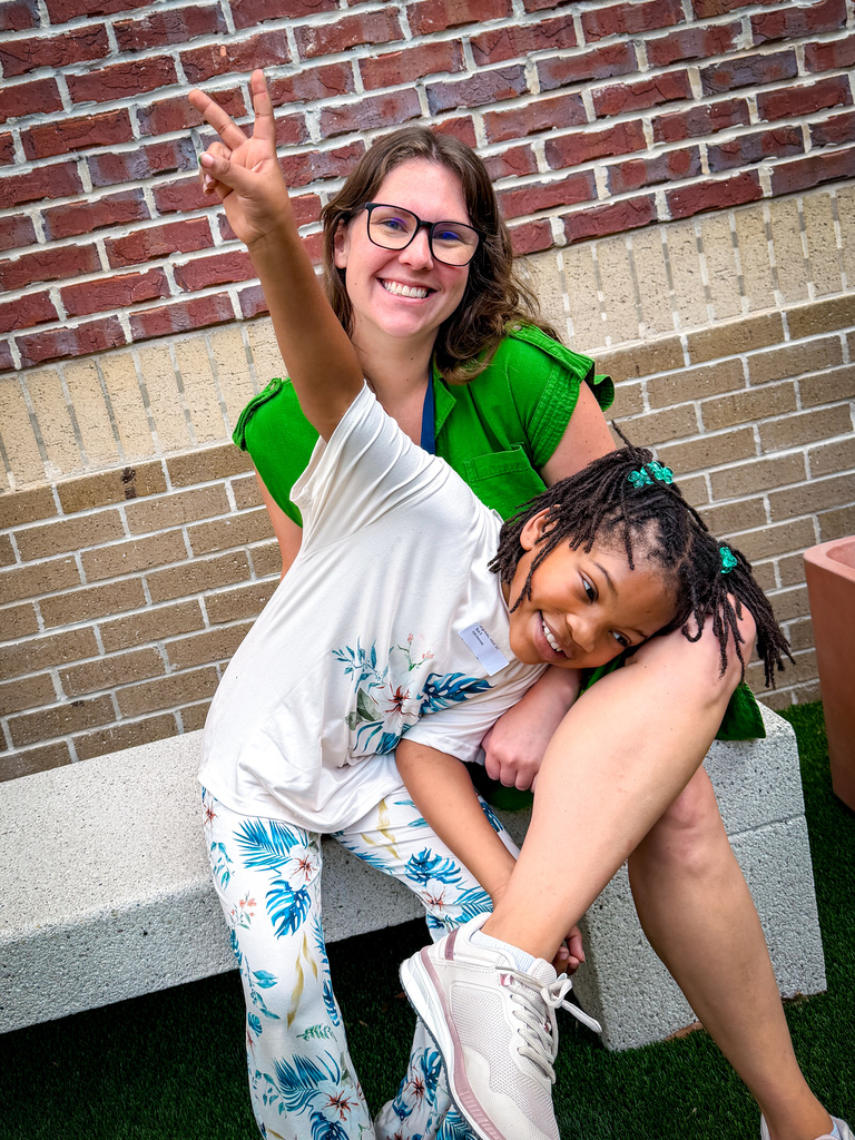 Woman in green holds a girl in white, seated on a bench. They pose with hands raised, smiling. Brick wall behind them.