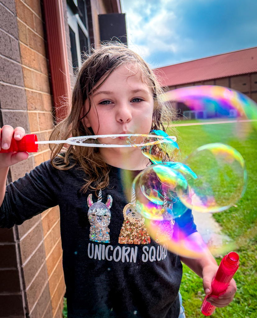 Girl in a black t-shirt with a unicorn image blows bubbles using a wand in a park.