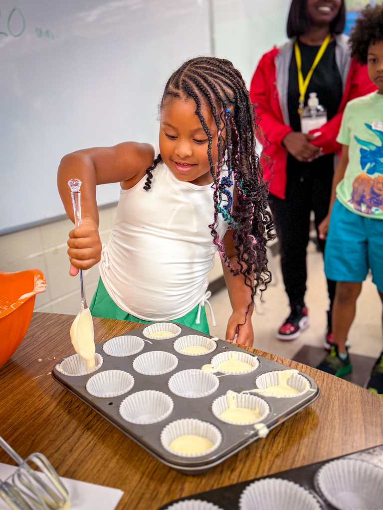A young girl adds ingredients to a muffin batter in a pan with two empty muffin tins nearby.