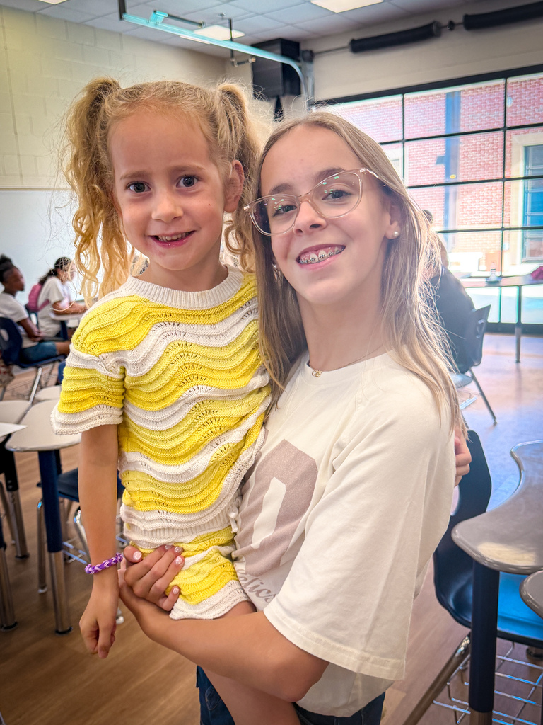 Two girls stand together indoors; one wears yellow stripes, the other has glasses and a white shirt. Behind them, chairs and desks are arranged in a classroom setting.