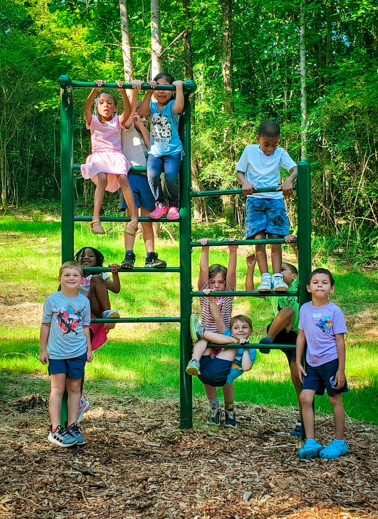 Group of children in various clothing and footwear, some hanging on a green monkey bar, on a grassy area with trees in background.