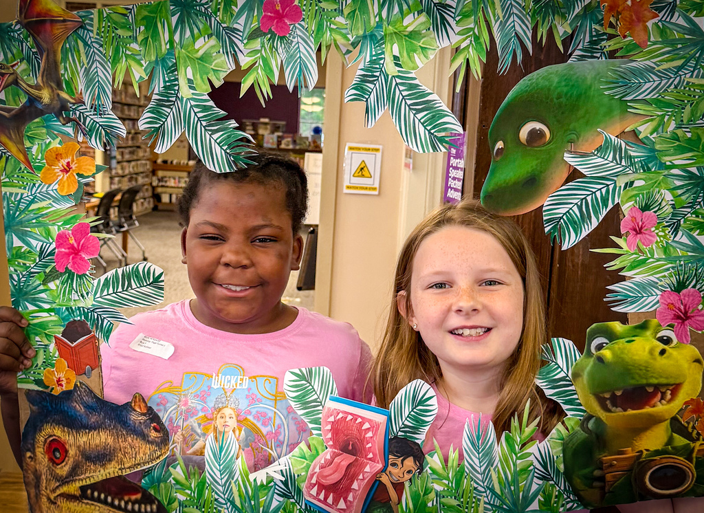 Two girls in pink shirts stand in a photo frame decorated with plants and animals.