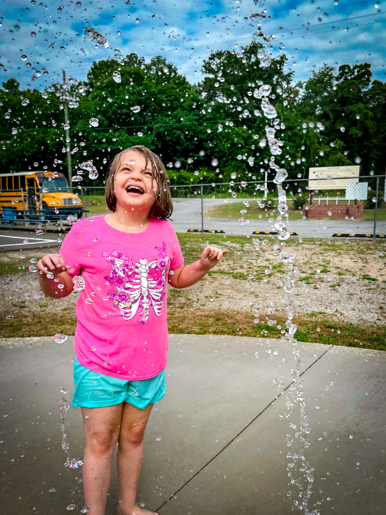 A young girl stands in water from a fountain, laughing with joy. She wears a pink shirt and green shorts.