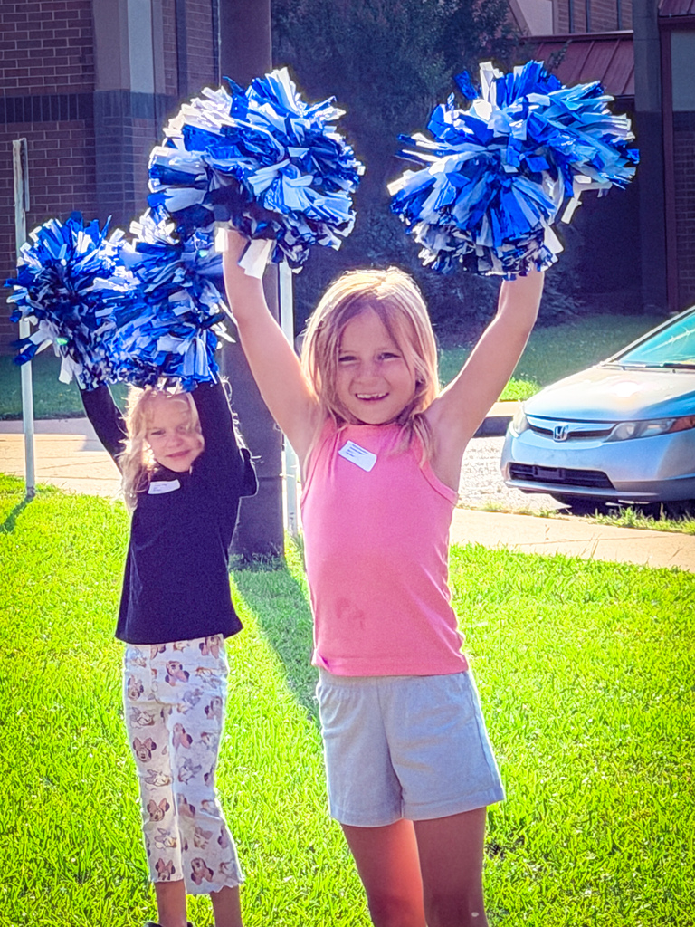 Two young girls holding blue and white pom poms in a grassy area with a building and car in the background.
