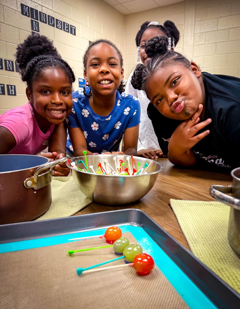Four girls with varying hairstyles, two wearing pink, one in blue, and one in black, smiling in a classroom.