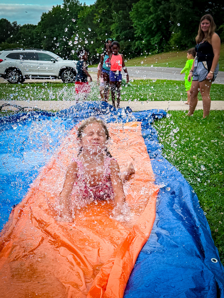 Girl slides down water-filled slide on grass. Other children and adults stand nearby. Trees and parked car visible in the background.