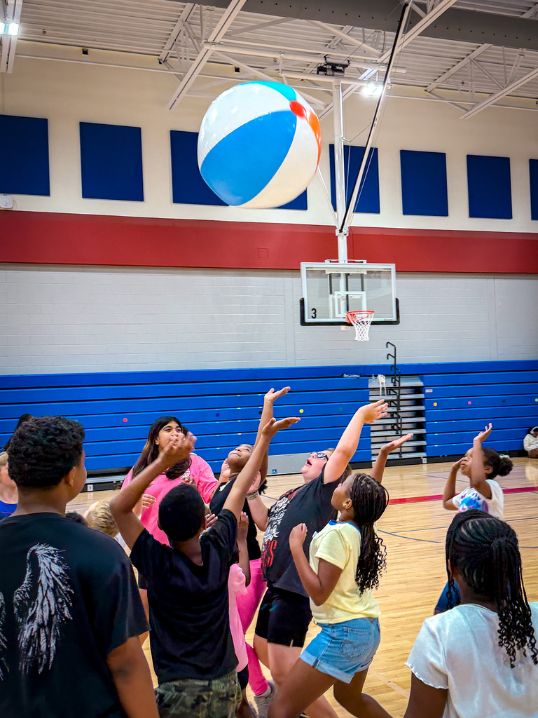 A group of children playing with a large beach ball in a gymnasium. Some are reaching up.