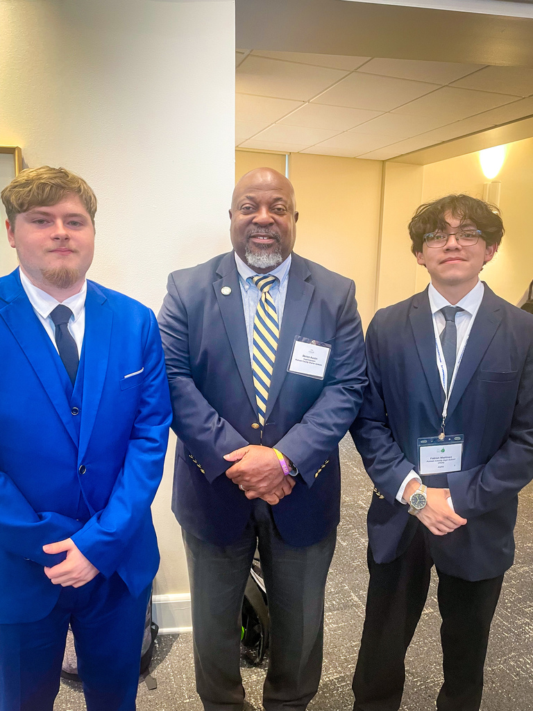 Three men in suits stand together, smiling, against a white wall. One wears a blue suit, another wears a dark suit and a yellow tie.