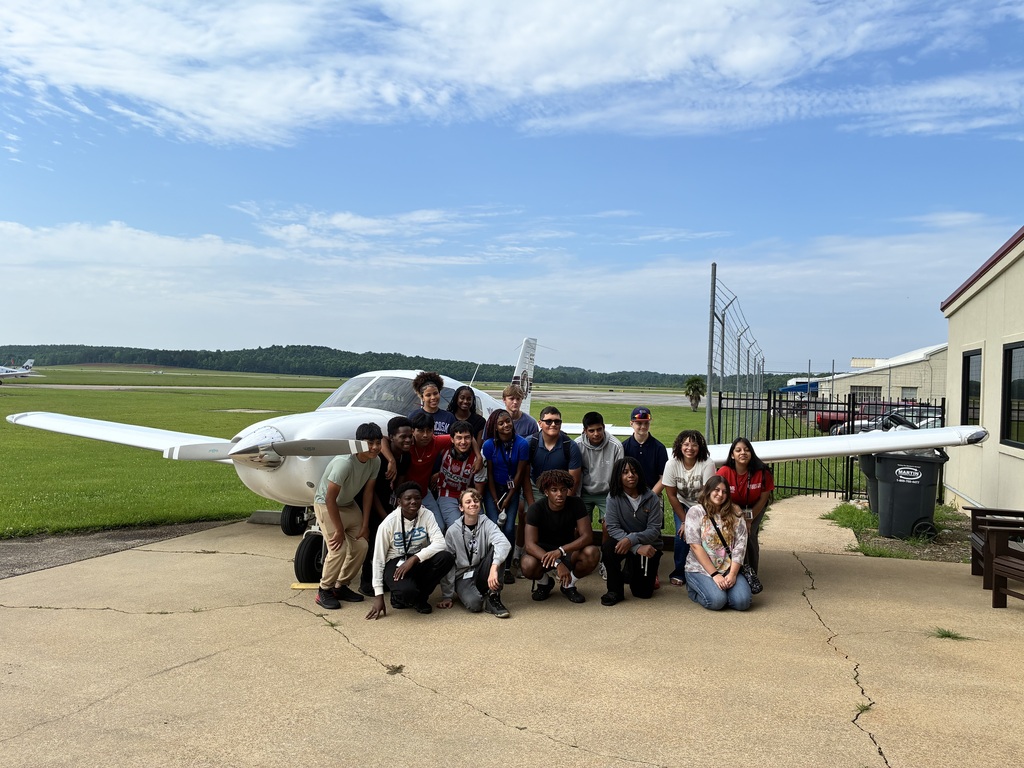 A group of people posing in front of a small white airplane, with a grassy area and a building in the background.