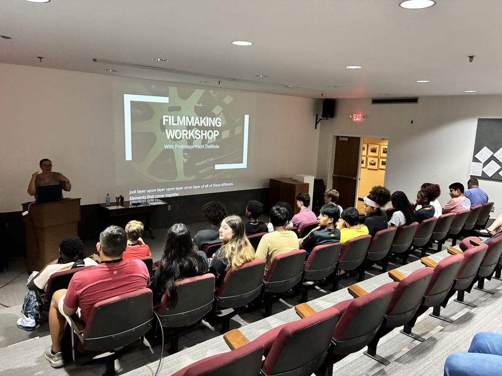 A classroom with rows of chairs and a person at a podium. A projector screen displays "FILMMAKING WORKSHOP."