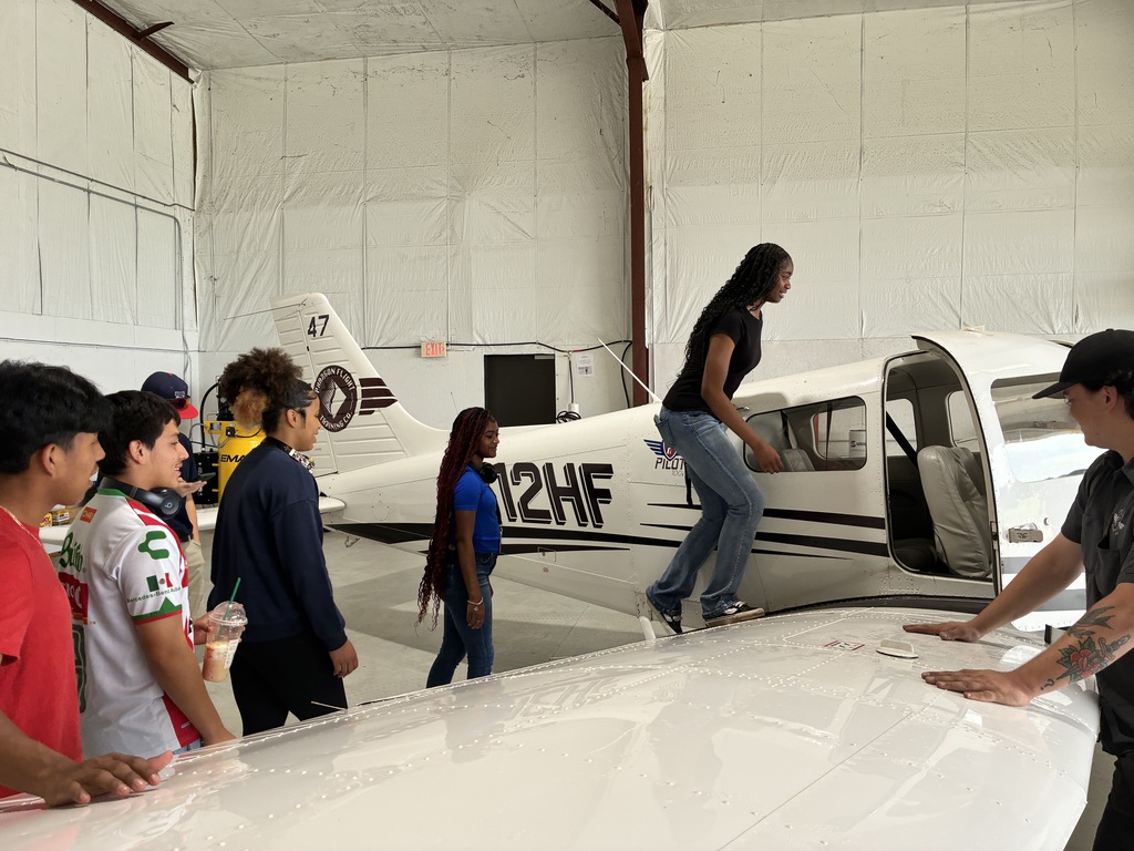 People gather near a white airplane in a hangar. A person stands on the wing while others watch.