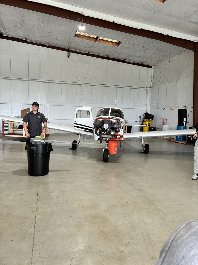 Two men in a hangar, one holding a trash bin. A small white plane is parked.