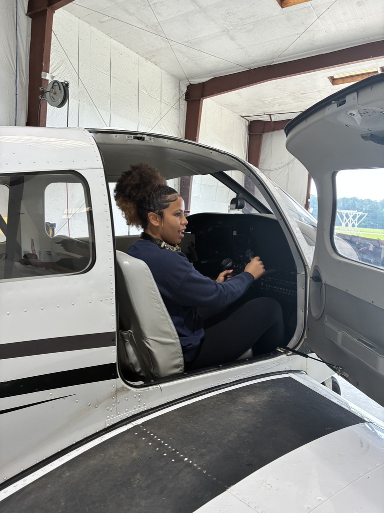 Two people are inside an open-door airplane, one in the cockpit and the other sitting in the cabin.
