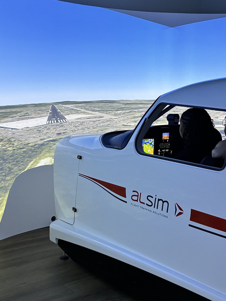 Cockpit of an aircraft simulator with a screen showing a flight path and runway. The aircraft's front features the word "alsim" and red stripes.