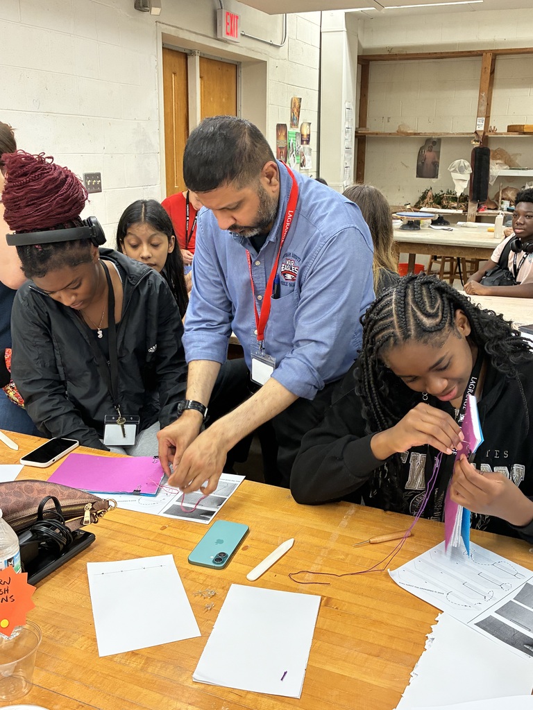 A teacher is assisting students with a project, working on a wooden table covered with papers and colorful objects.