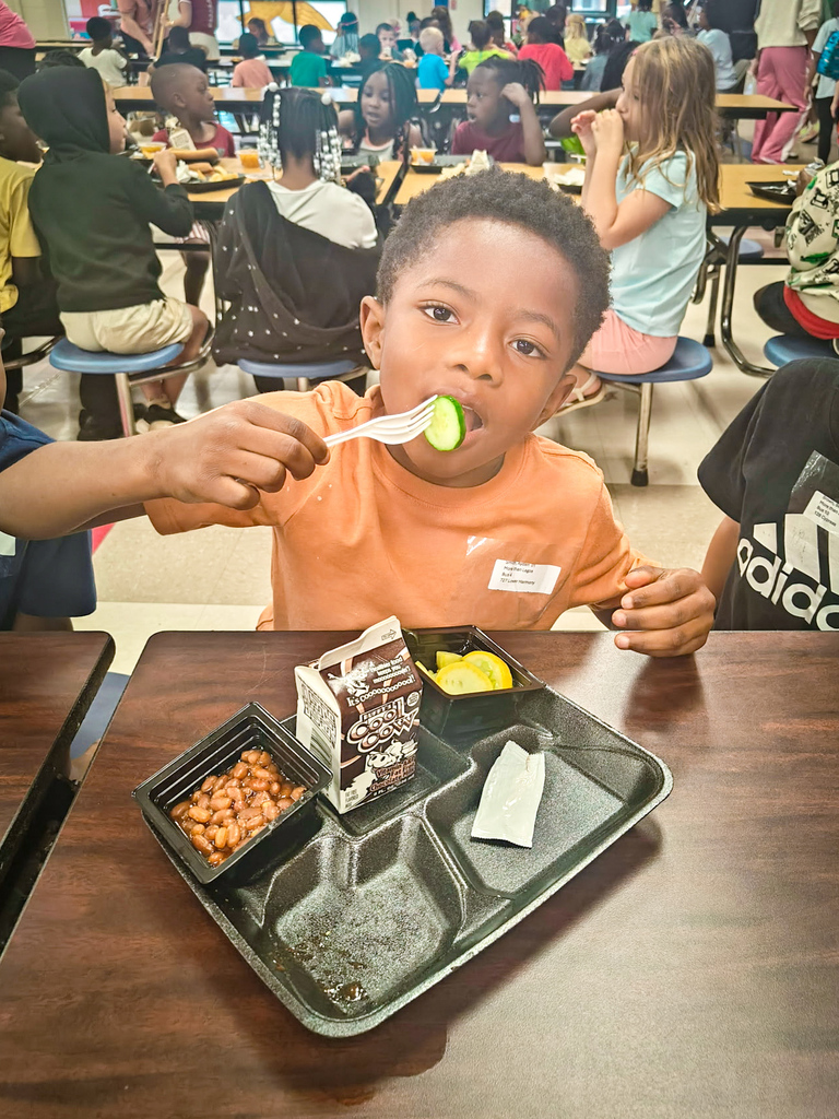 Child in orange shirt eating food from a cafeteria tray with food and a juice carton.