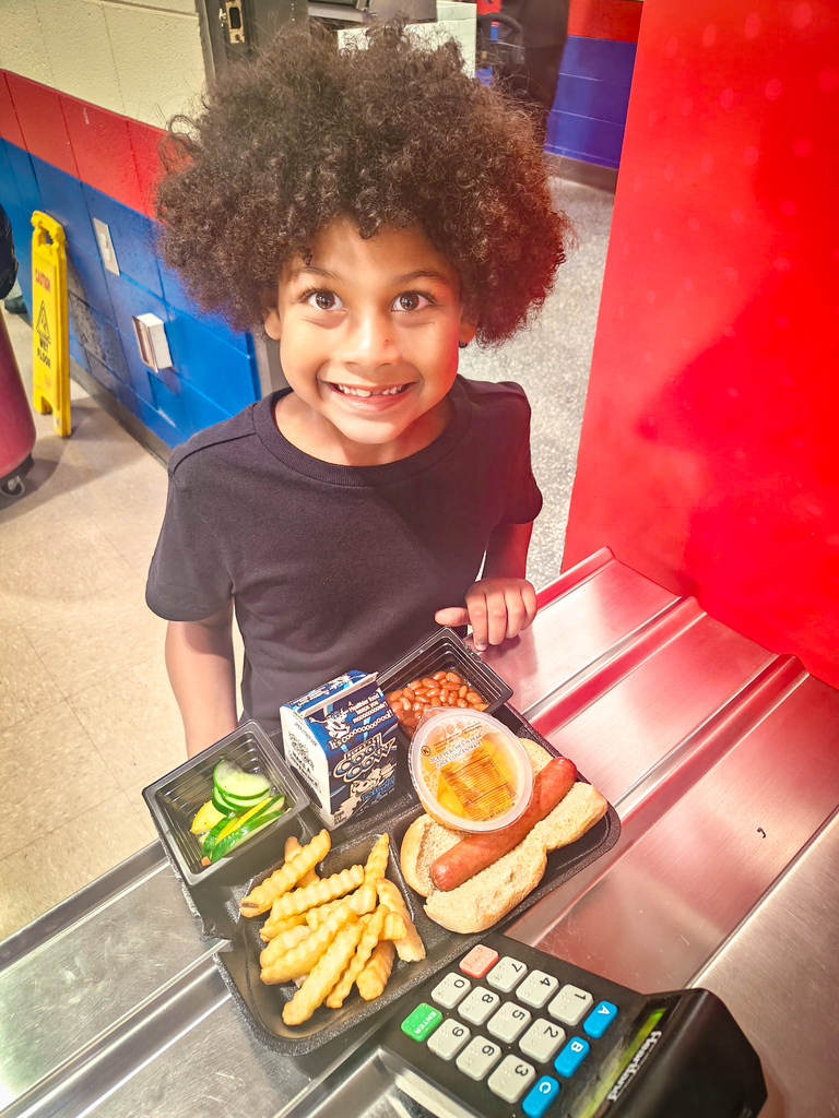 A smiling child with curly hair holds a lunch tray with food items. Behind, a red wall and a yellow sign.