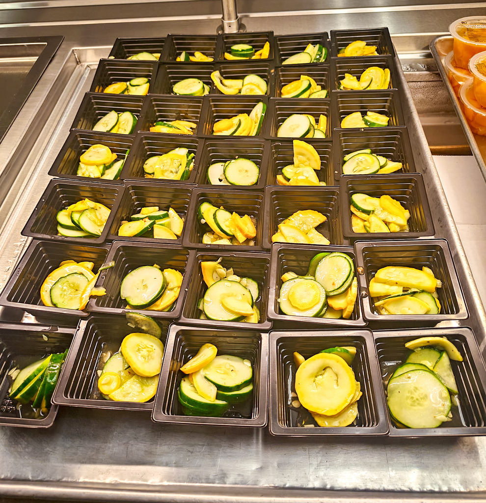 Many black plastic containers with sliced yellow zucchini on a metal tray. Containers are in rows.