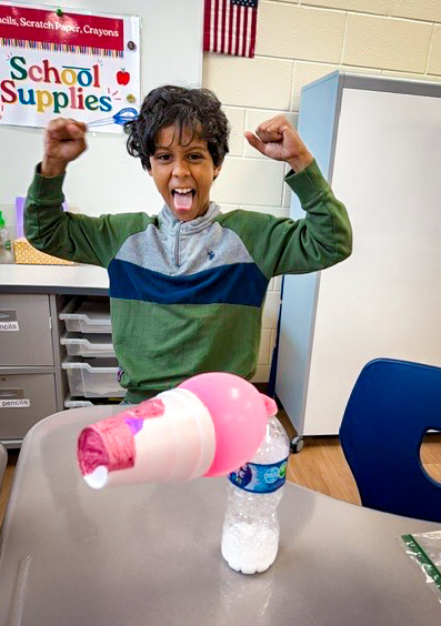 A young person with arms raised inside a classroom, holding a pink object over a plastic bottle.