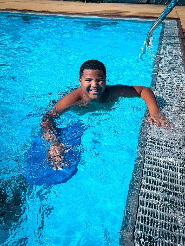Person in pool holding blue object, smiling, and splashing water while on the edge of the pool.